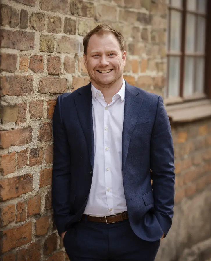 Press photo 02 of David Jacoby: Smiling, dark blazer, brick wall